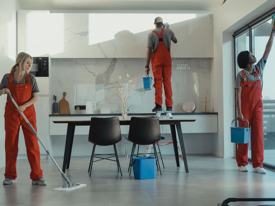 Three cleaners in orange overalls mopping the floor and wiping surfaces in a modern kitchen and dining area