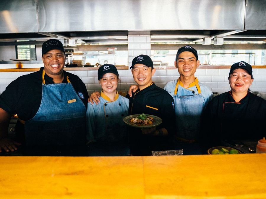 Five smiling kitchen staff in chef jackets and aprons behind a restaurant counter, one holding a plated dish