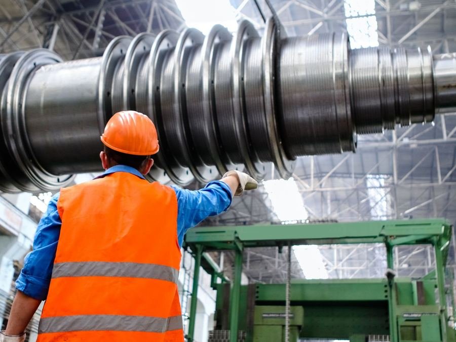 A factory worker in an orange hard hat and high-visibility vest pointing at large industrial machinery