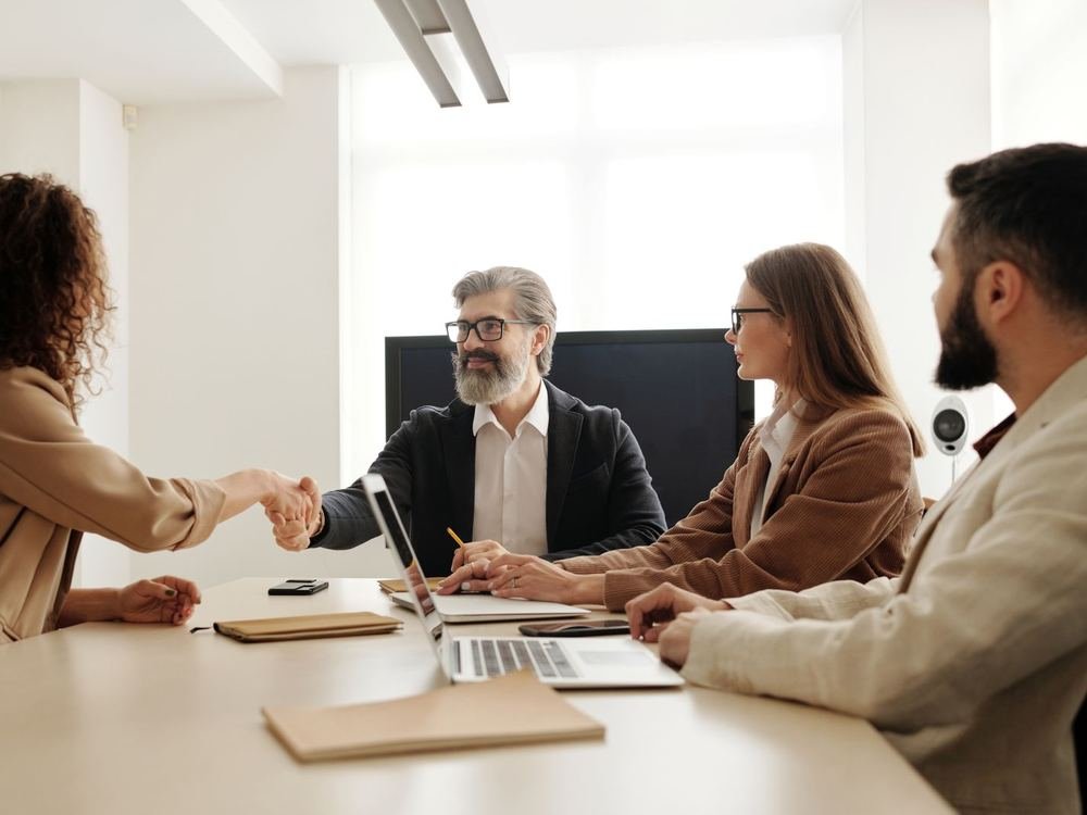 A business meeting in which a woman shakes hands with a bearded man across a table while two colleagues look on